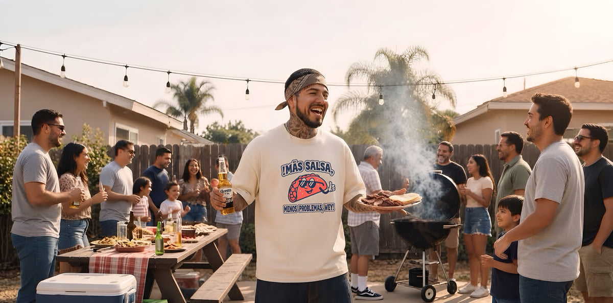 Mexican American man at a backyard family BBQ wearing a cream ¡OYE! tee that says “Más salsa, menos problemas, wey,” holding a beer and a plate of carne asada and tortillas.